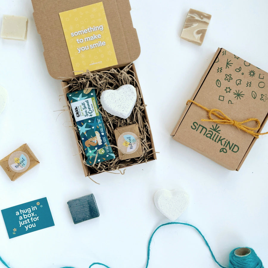 Deluxe Hug in a Box Gift set including oat milk salted caramel chocolate bar, natural heart shaped bath bomb, mini face wash bar. The products are inside a kraft box nestled in shredded paper and tied with twine. 