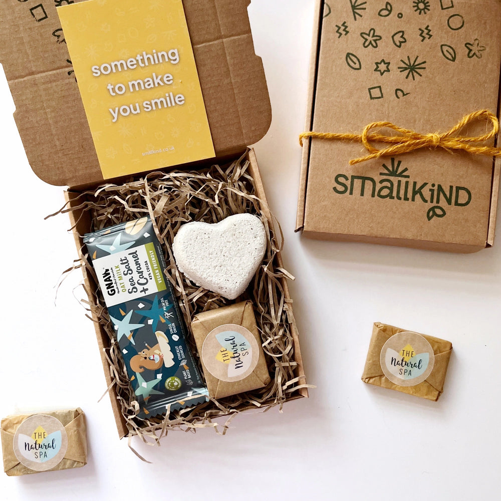 Deluxe Hug in a Box Gift set including oat milk salted caramel chocolate bar, natural heart shaped bath bomb, mini face wash bar. The products are inside a kraft box nestled in shredded paper and tied with twine. 