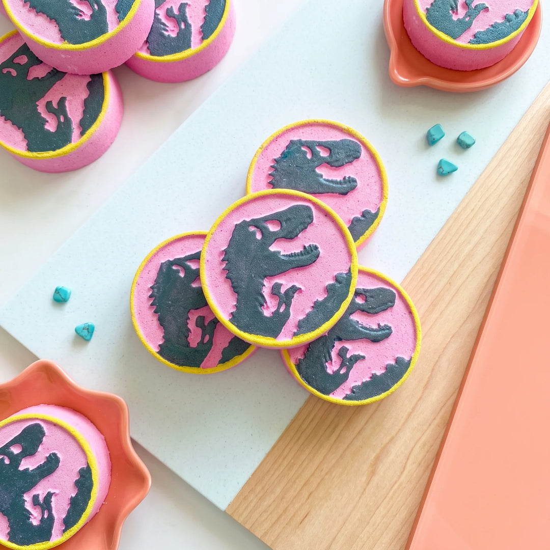 a collection of pink circular bath bombs with dinosaur silhouettes printed on them and a yellow border. 
