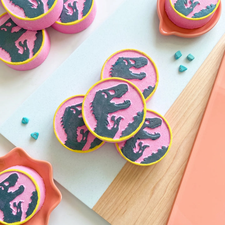 a collection of pink circular bath bombs with dinosaur silhouettes printed on them and a yellow border. 