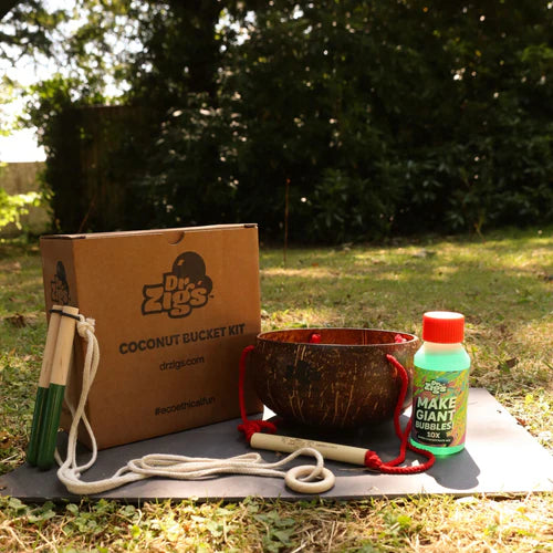 children's bubble kit with coconut bucket, bottle of concentrated bubble mix, giant wooden wand and a gift box. The items are laid out on a garden lawn. 
