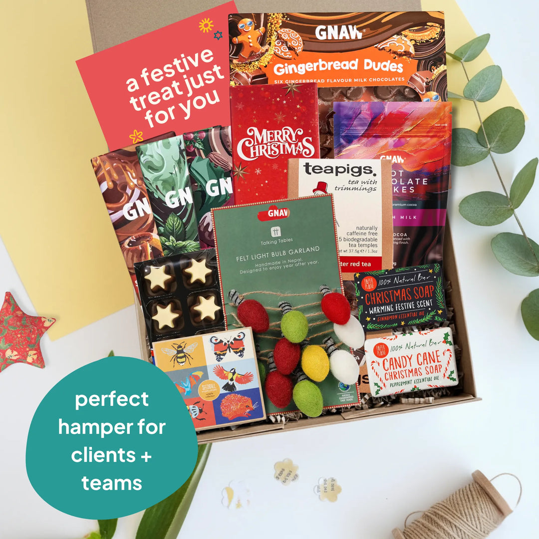 A curated Christmas gift box filled with assorted treats and packaged snacks, photographed from above on a white surface with decorative props. Visible items include GNAW gingerbread cookies, a red &