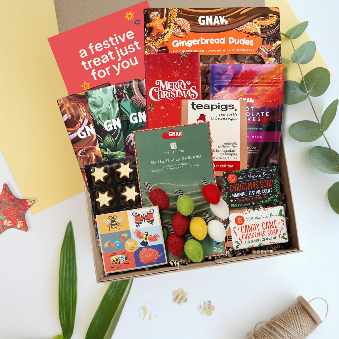 A curated gift box filled with assorted packaged treats and snacks, including GNAW gingerbread cookies, a Merry Christmas tea bag, a festive hair garland, candy canes, and a red greeting card that reads &