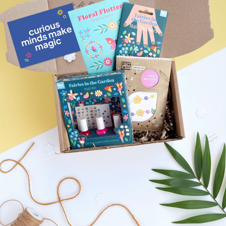 Children's craft kit with fairy and flower themed nail kit, tattoos and jewellery making set and a 'curious minds make magic' card on a white surface with green leaves.