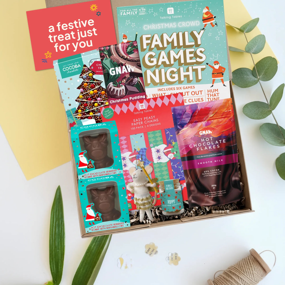 A Christmas-themed family game night gift box featuring a teal and red 'Family Games Night' box, chocolate bars, paper chains craft kit, mouse Christmas decoration, hot chocolate flakes and a jar of seedballs styled on a white surface with decorative leaves and a yellow card.