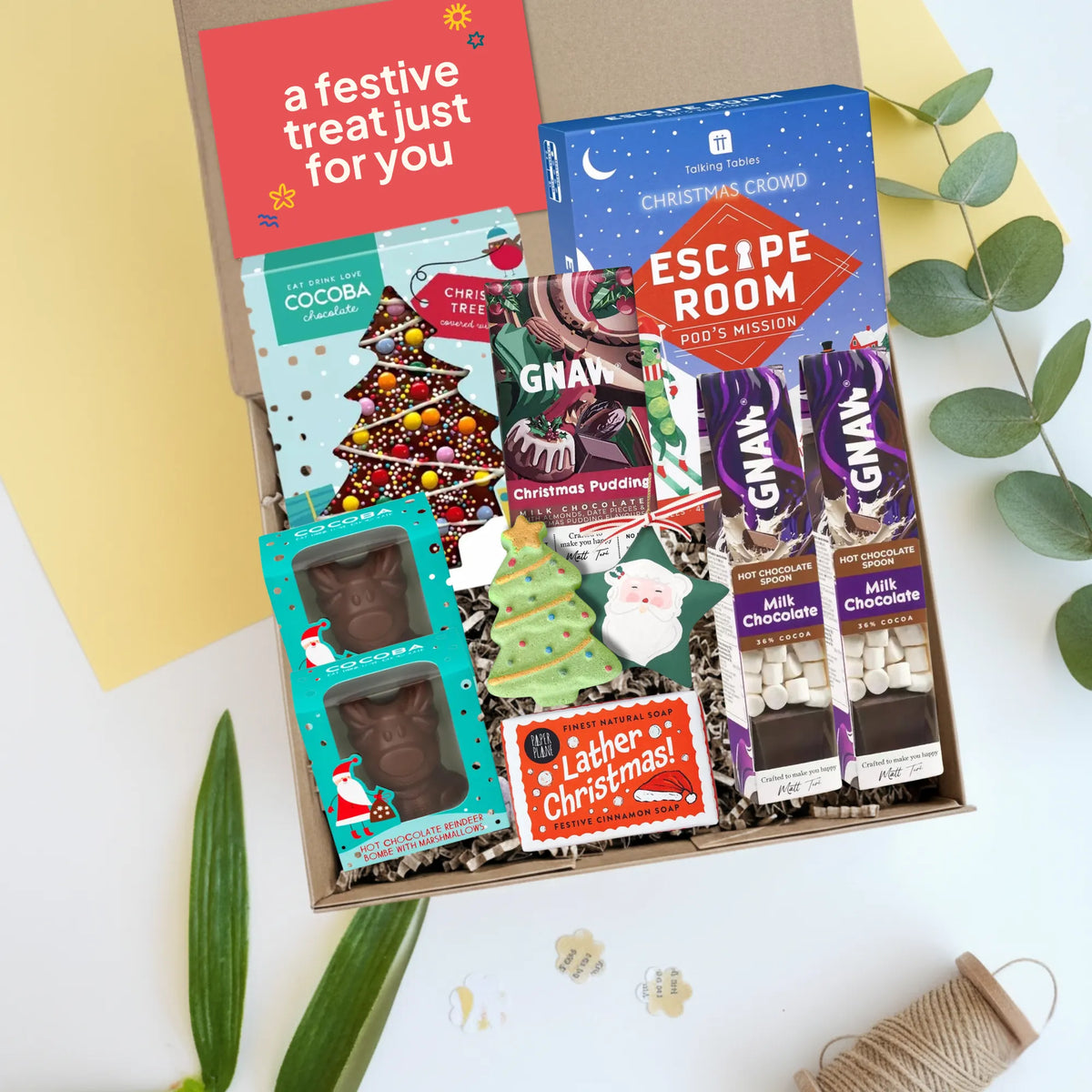 A curated Christmas gift box filled with assorted treats and packaged snacks, photographed from above on a white surface with decorative leaves and paper. Visible items include a red card reading 'a festive treat just for you,' a blue 'Escape Room Mission' game, a teal 'Cocoba' chocolate bombs, Christmas Pudding chocolate bar,  Ganw hot chocolate stirrers, lather Christmas soap bar, christmas tree bath bomb and a green tree ornament. 