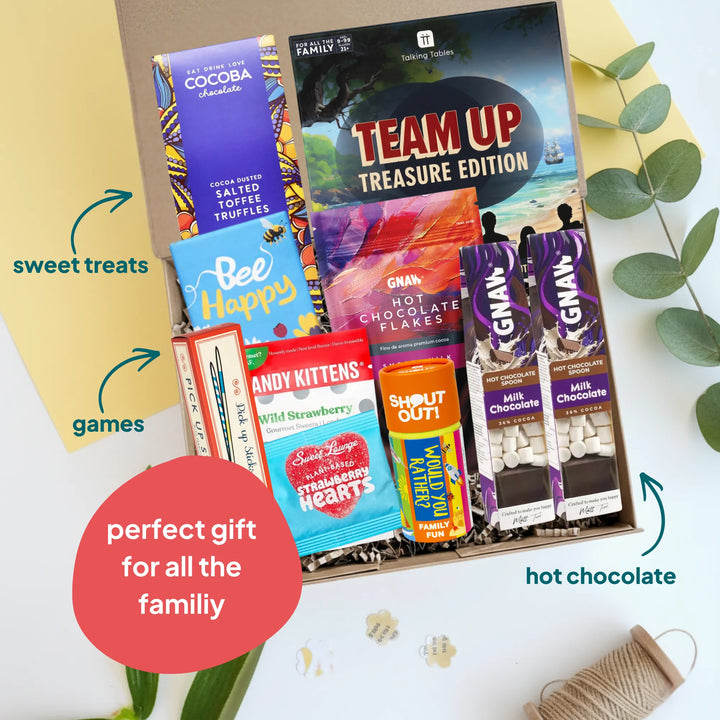 A curated gift box filled with assorted snack items and a board game, photographed from above on a white surface with decorative leaves and paper. Visible products include a Team Up Treasure Edition game, a blue Bee Happy chocolate bar, a red and blue Candy Kittens Wild Strawberry sweets, a yellow Shout Out! game, pick up sticks game and a selection on Gnaw chocolates.
