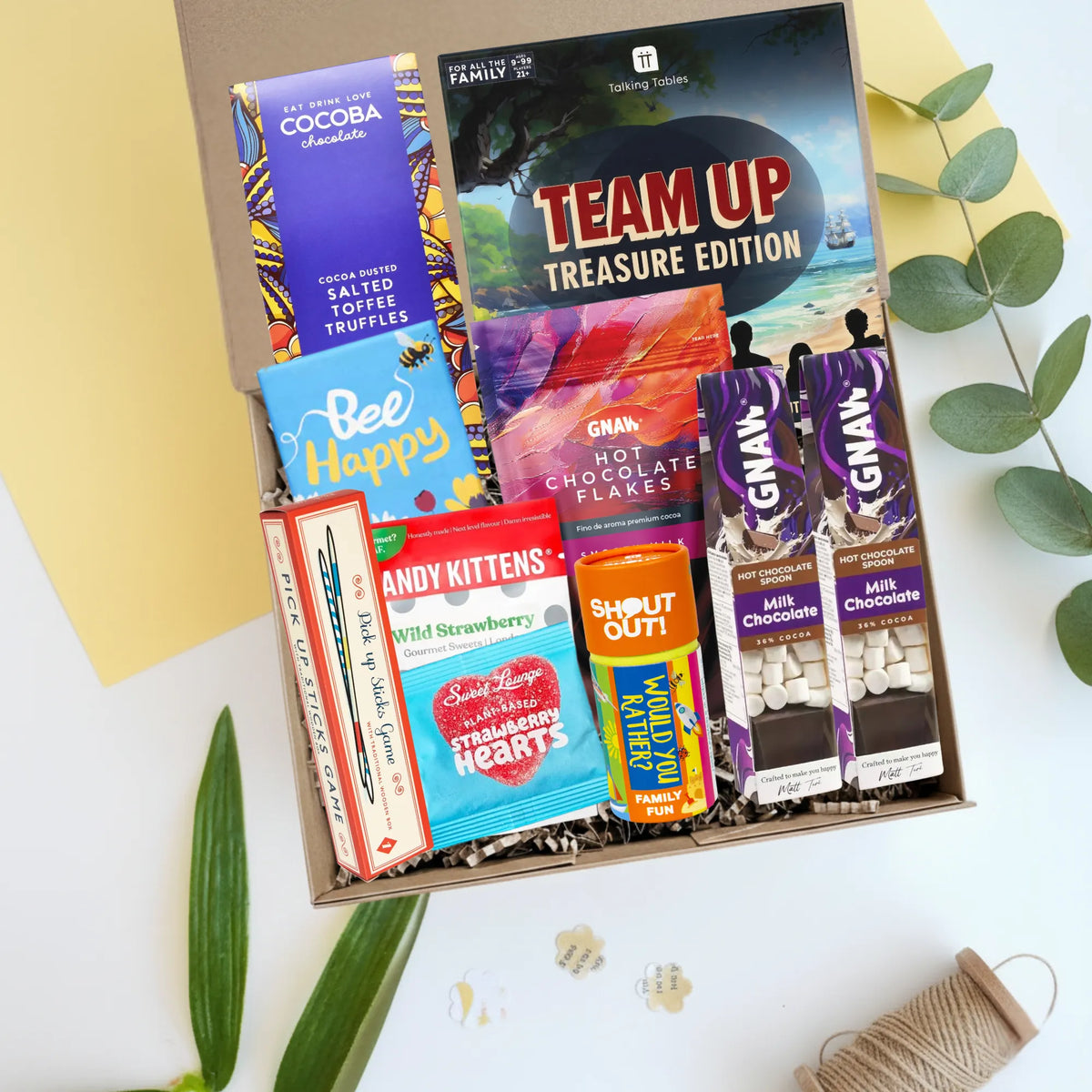 A curated gift box filled with assorted snack items and a book, photographed from above on a white surface with decorative leaves and paper. Visible products include a Team Up Treasure Edition book, a blue Bee Happy chocolate bar, a red and blue Candy Kittens Wild Strawberry sweets, a yellow Shout Out! game, pick up sticks game and a selection on Gnaw chocolates.