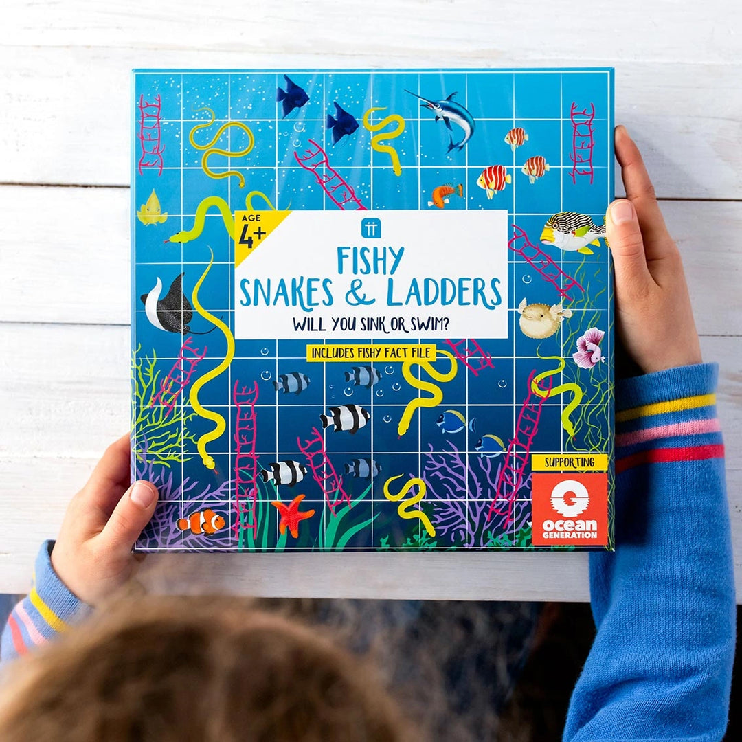 Child holding a 'Fishy Snakes & Ladders' board game box with ocean-themed illustrations.