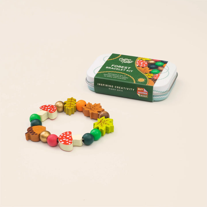 A forest-themed bead bracelet kit with colorful wooden beads shaped like trees, mushrooms, acorns, and leaves, shown next to its green and white tin packaging on a light beige background.