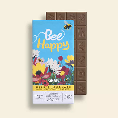 'Bee Happy' Honeycomb + Caramel Milk Chocolate Bar in colourful packaging with illustrations of flowers and bees. A bar of Gnaw milk chocolate is in the background.