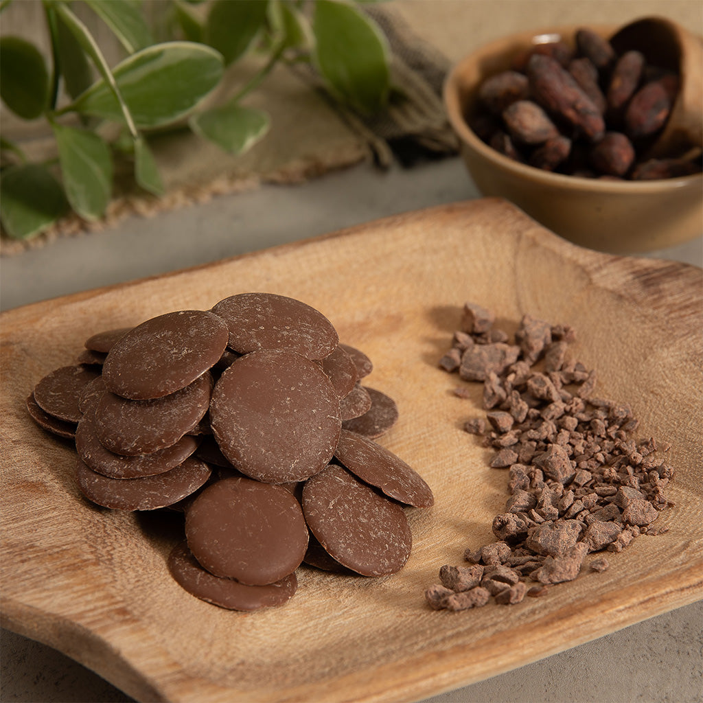 Chocolate buttons and cocoa powder on a wooden plate with a bowl of cocoa beans in the background.