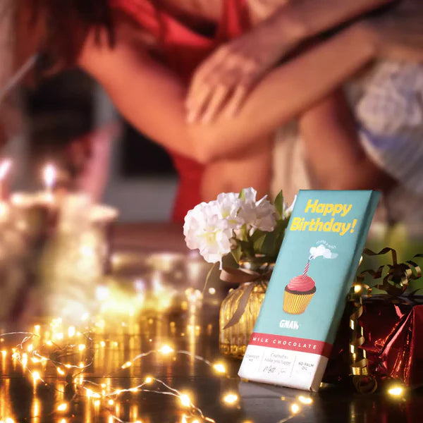 Happy Birthday Milk Chocolate Bar with a blue wrapper featuring a cupcake and happy birthday message. The chocolate is on a  table with fairy lights, gifts and flowers.
