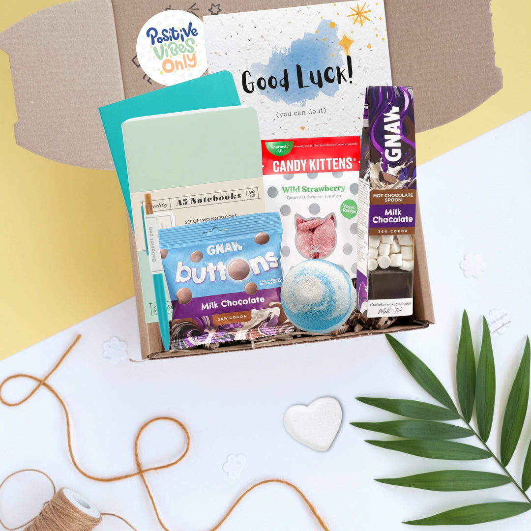 Gift box including Greeting card with 'Good Luck!' message, chocolate bars, stationery, bath bomb and other items in a box on a decorative background.