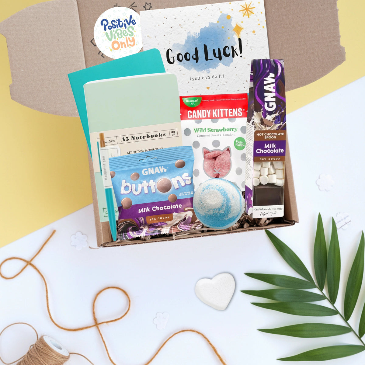 Gift box including Greeting card with 'Good Luck!' message, chocolate bars, stationery, bath bomb and other items in a box on a decorative background.