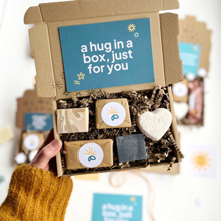 Pamper Gift Box of small size solid soap bars and toiletries with a heart-shaped bath bomb nestled in shredded paper. The box has a 'hug in a box just for you card inside and is held by a person wearing a mustard yellow sweater.