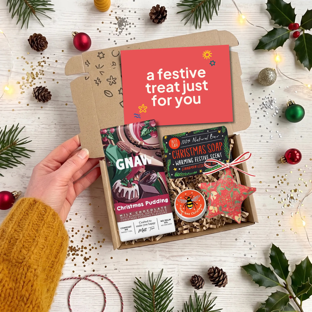 A hand holds open a kraft gift box filled with Christmas-themed treats on a white wooden surface decorated with pine branches, ornaments, fairy lights, and confetti. The box contains a red card reading 'a festive treat just for you,' a packaged chocolate pudding chocolate bar by GNAW, a Christmas soap bar, and a red star-shaped ornament.