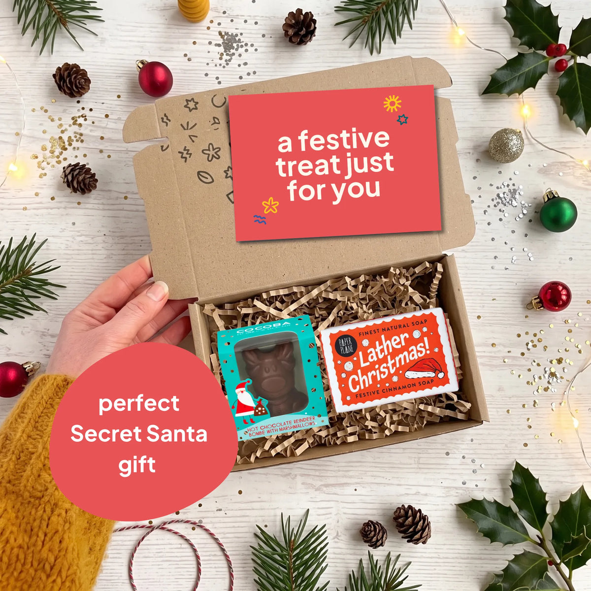 A hand holds open a brown cardboard gift box filled with shredded paper, containing a chocolate reindeer figure in a blue box and a  'Lather Christmas!' cinnamon soap bar. The scene is styled with Christmas greenery, ornaments, fairy lights, and a red card reading 'a festive treat just for you' and 'perfect Secret Santa gift'.