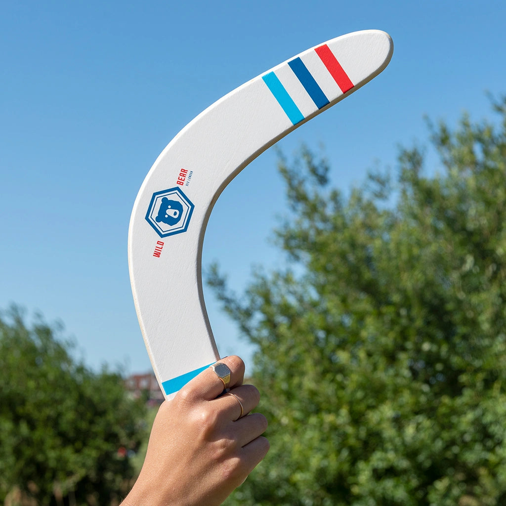 hand holding up a Rex London Wild Bear Wooden Boomerang with blue and red retro style design. Trees and sky are in the background. 