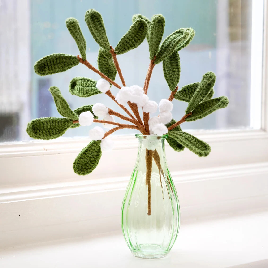 Crocheted mistletoe - green leaves with white berries in a clear glass vase on a windowsill.