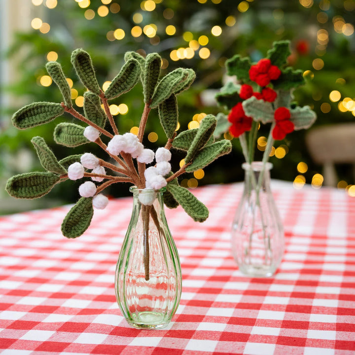 Handmade crochet mistletoe branches with green leaves and white berries in a small clear glass vase, set on a red and white checkered tablecloth with a blurred Christmas tree and lights in the background.