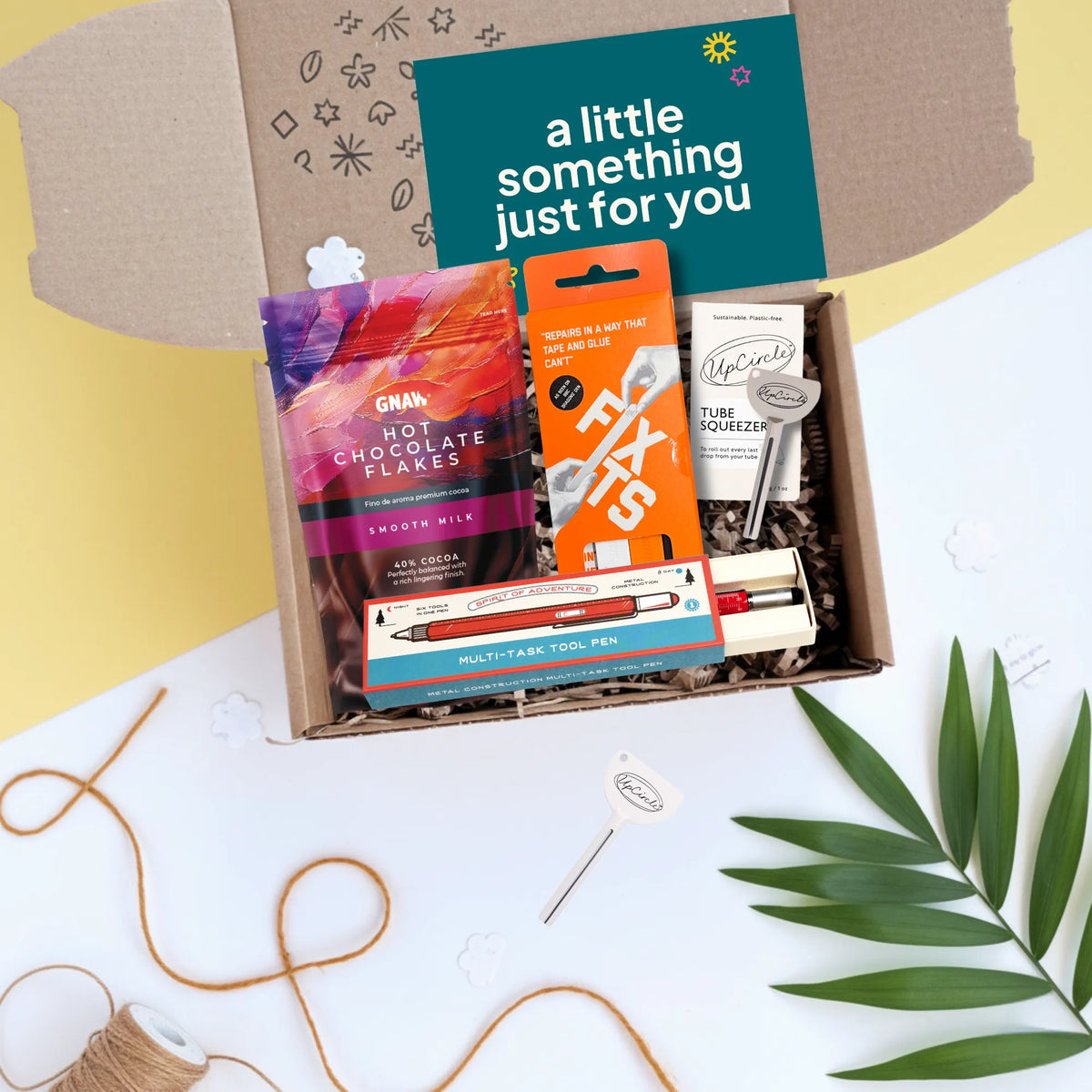 An open cardboard gift box filled with assorted packaged snacks and stationery, photographed from above on a white surface with decorative twine, leaves, and a card reading 'a little something just for you.' Visible items include a packet of GNAW hot chocolate flakes, an orange FIXTIS pack, handy tube squeezer and a multicolour multi-tool pen.