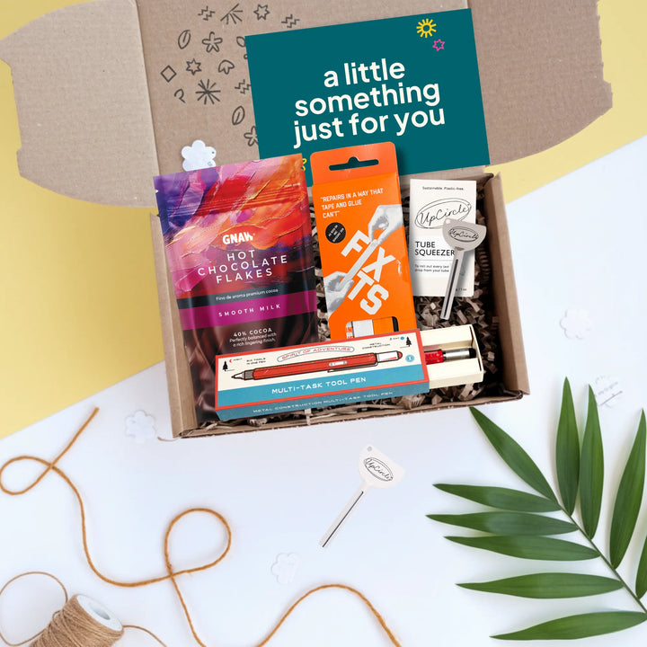 An open cardboard gift box filled with assorted packaged snacks and stationery, photographed from above on a white surface with decorative twine, leaves, and a card reading &
