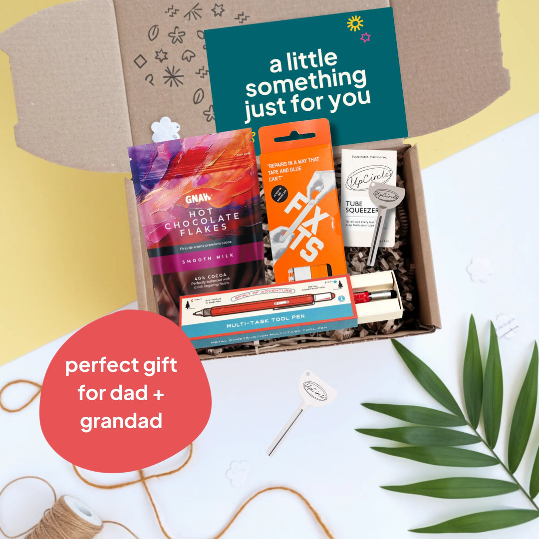 An open gift box arranged on a white surface with a yellow side background. Inside the box are a packet of GNAW hot chocolate flakes, a box of Fixtis tools, a tube squeezer, and a multi tool pen.