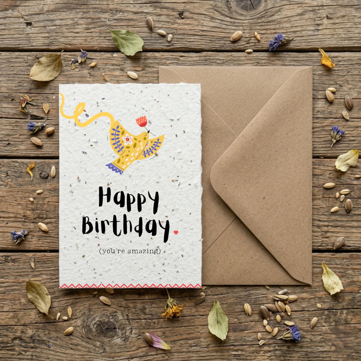 happy birthday card with a yellow folk inspired bird design and the words 'happy birthday (you're amazing) in a handwritten style font. The card is laid on top of a brown envelope on a wooden table.