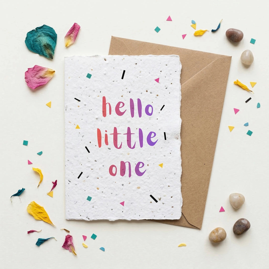 Greeting card with 'hello little one' text on a textured paper with a brown envelope, surrounded by colorful confetti and small stones.