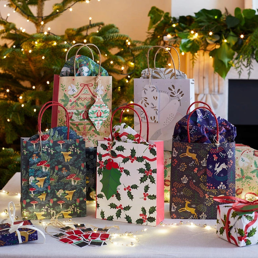 Collection of Christmas-themed gift bags and a box on a table with a decorated tree in the background.