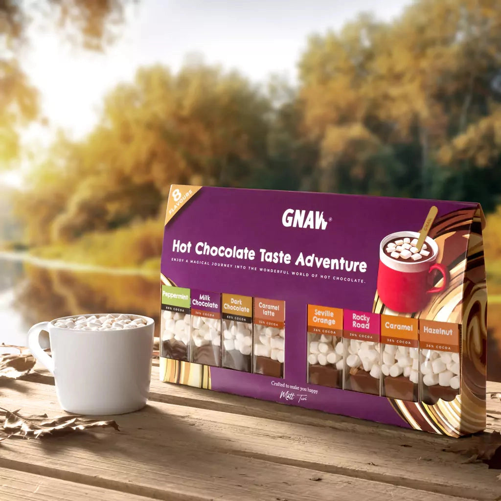 A bag of GNAW Hot Chocolate Taste Adventure mix sits on a wooden table next to a white mug filled with hot chocolate and marshmallows, with a blurred autumn forest background.