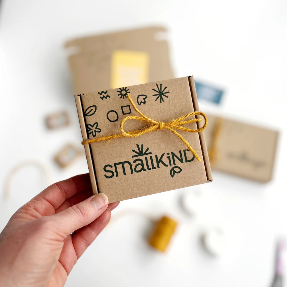 white woman's hand holding a Smallkind Hug in a box gift set in a stamped kraft box tied with mustard yellow twine