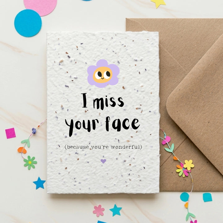 Greeting card with 'I miss your face (because you're wonderful)' text and a flower illustration, accompanied by a brown envelope on a decorative background.