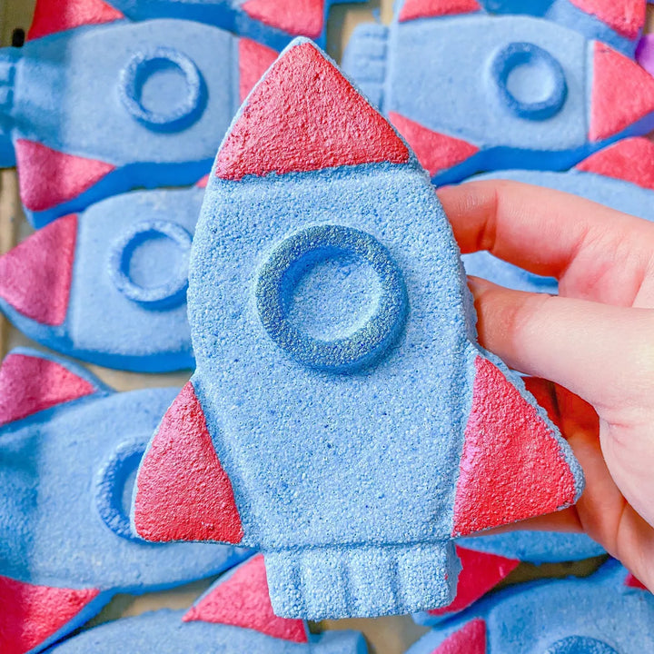 Blue and red rocket-shaped bath bombs held by a hand with more bath bombs in the background.
