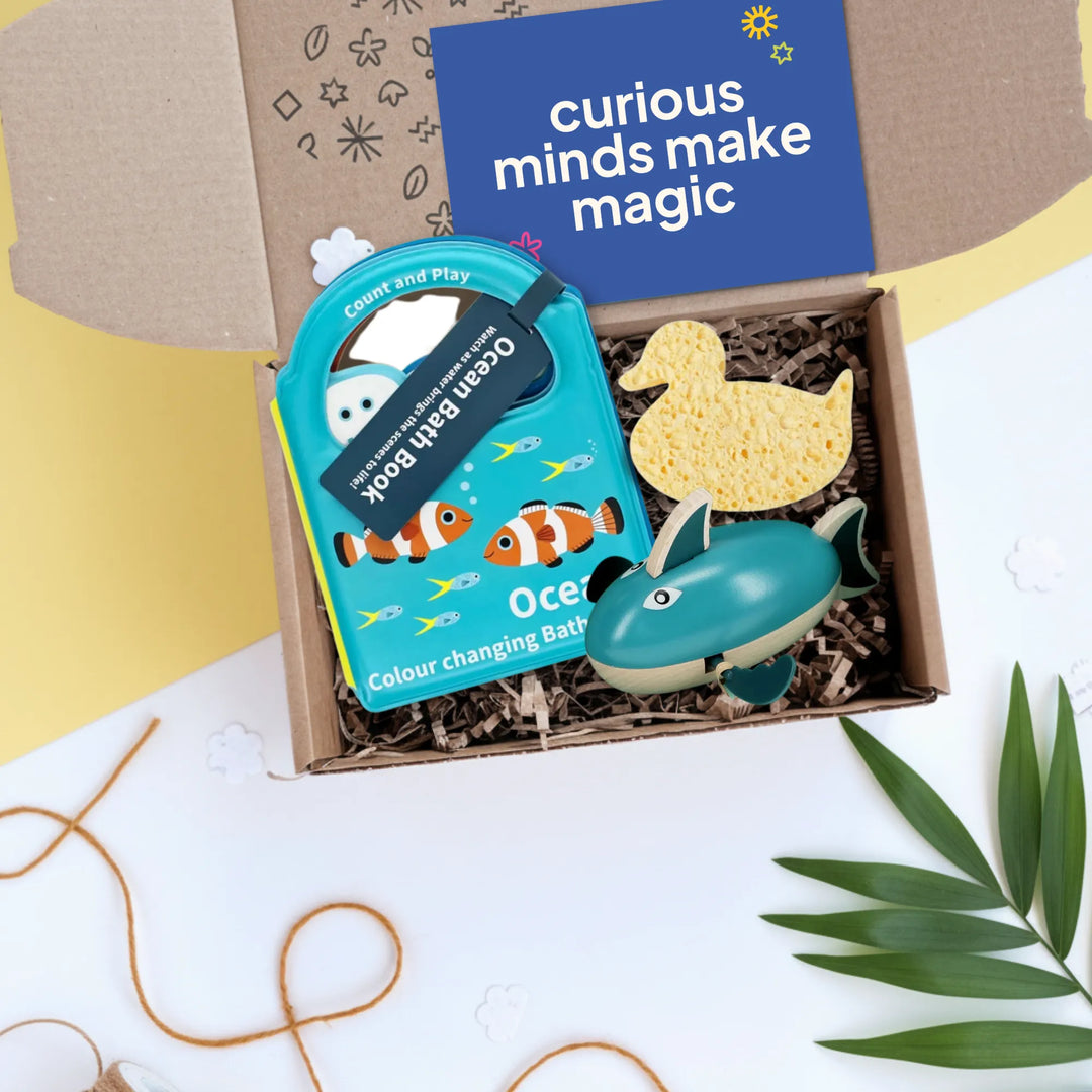 An open cardboard gift box filled with bath and bath-toy items, photographed from above on a white surface with a pale yellow background. Visible contents include a blue book titled &