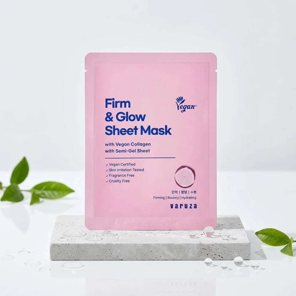 A pink Varuza Firm &amp; Glow Sheet Mask pouch standing upright on a light gray stone slab with decorative green leaves around it, against a clean white background.