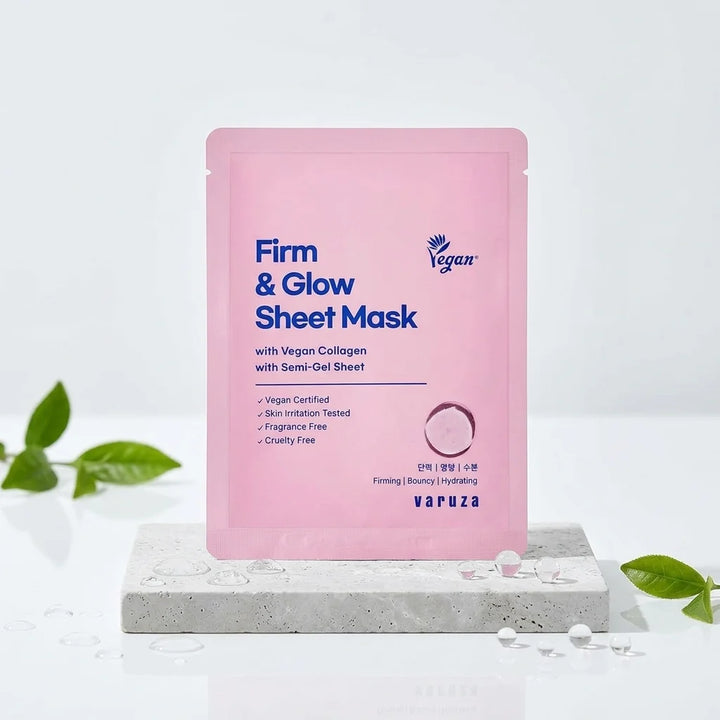 A pink Varuza Firm &amp; Glow Sheet Mask pouch standing upright on a light gray stone slab with decorative green leaves around it, against a clean white background.