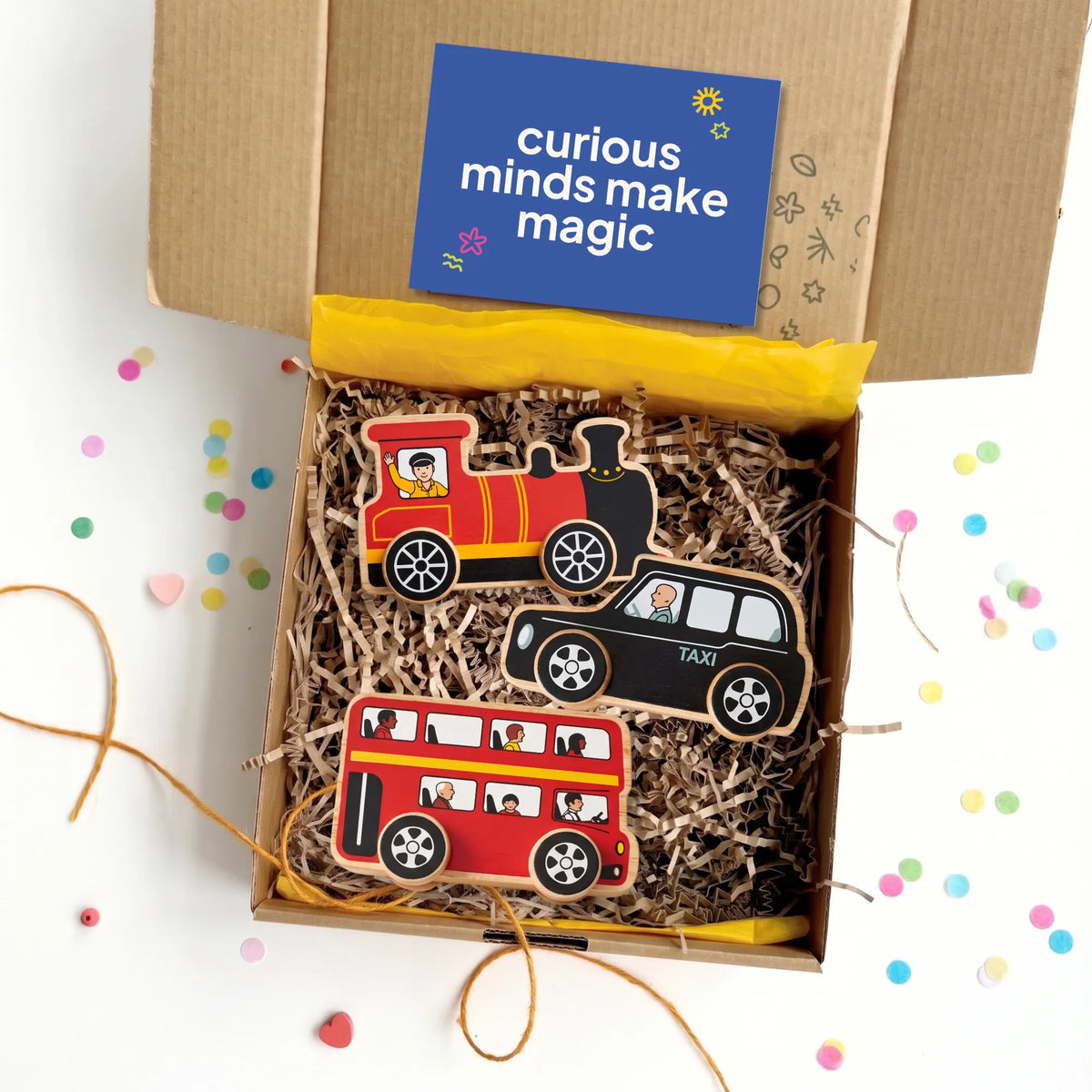 A gift box containing three wooden train-themed cookies: a red steam locomotive, a black taxi, and a red double-decker bus, arranged in shredded paper inside a brown box. A blue card reading 'curious minds make magic' is visible at the top, with confetti scattered on the white surface around the box.
