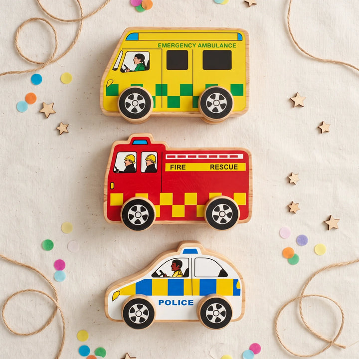 Three wooden toy vehicles for children: an emergency ambulance, a fire rescue truck, and a police car, arranged vertically on a light beige fabric background with decorative twine and confetti.