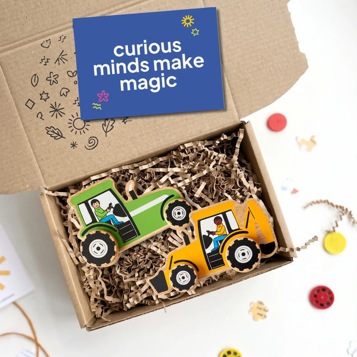 An open cardboard gift box filled with brown paper shred, holding two wooden toy tractors in green and yellow, with a blue card reading &