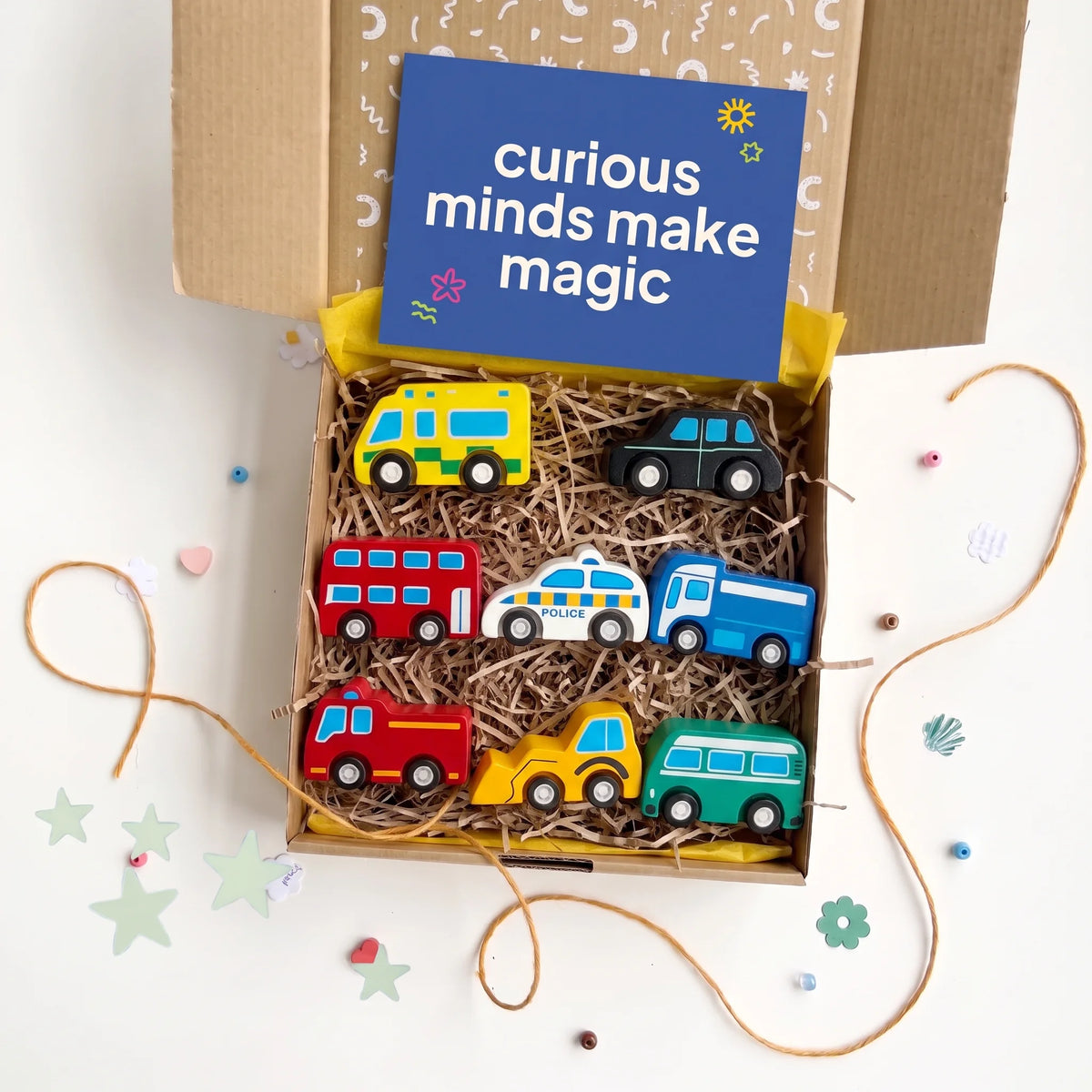 An open cardboard gift box filled with shredded paper and six colorful wooden toy vehicles, including buses, cars, and a truck, with a blue card reading 'curious minds make magic' inside. The box is photographed on a white surface with decorative paper stars and twine around it.