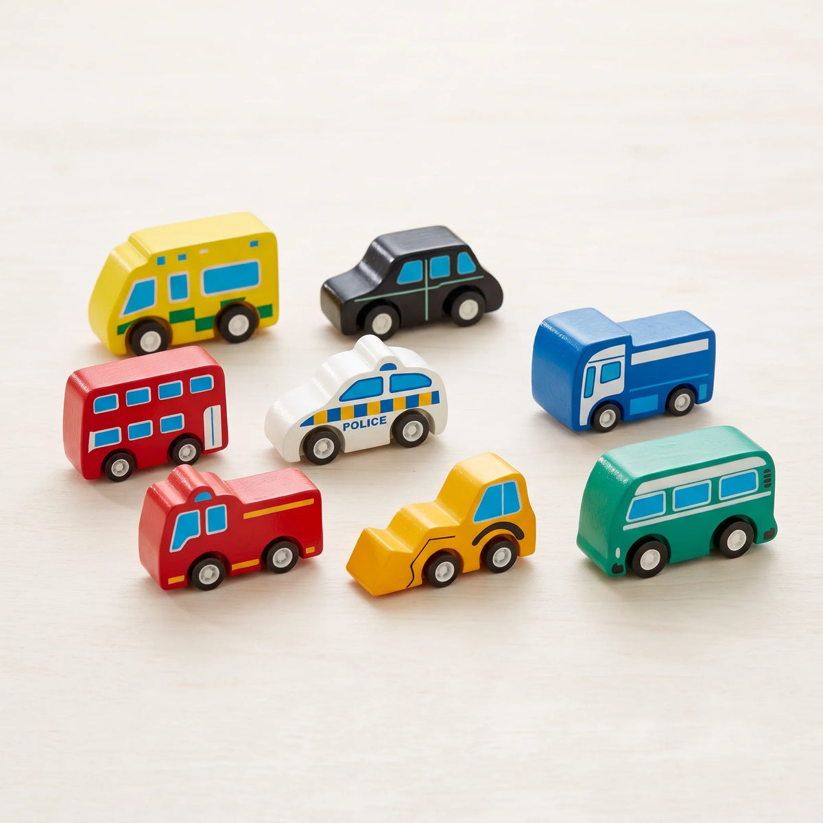 A set of colourful wooden toy vehicles, including buses, a car, a police car, a fire truck, a construction vehicle, and a van, arranged on a light beige wooden surface.