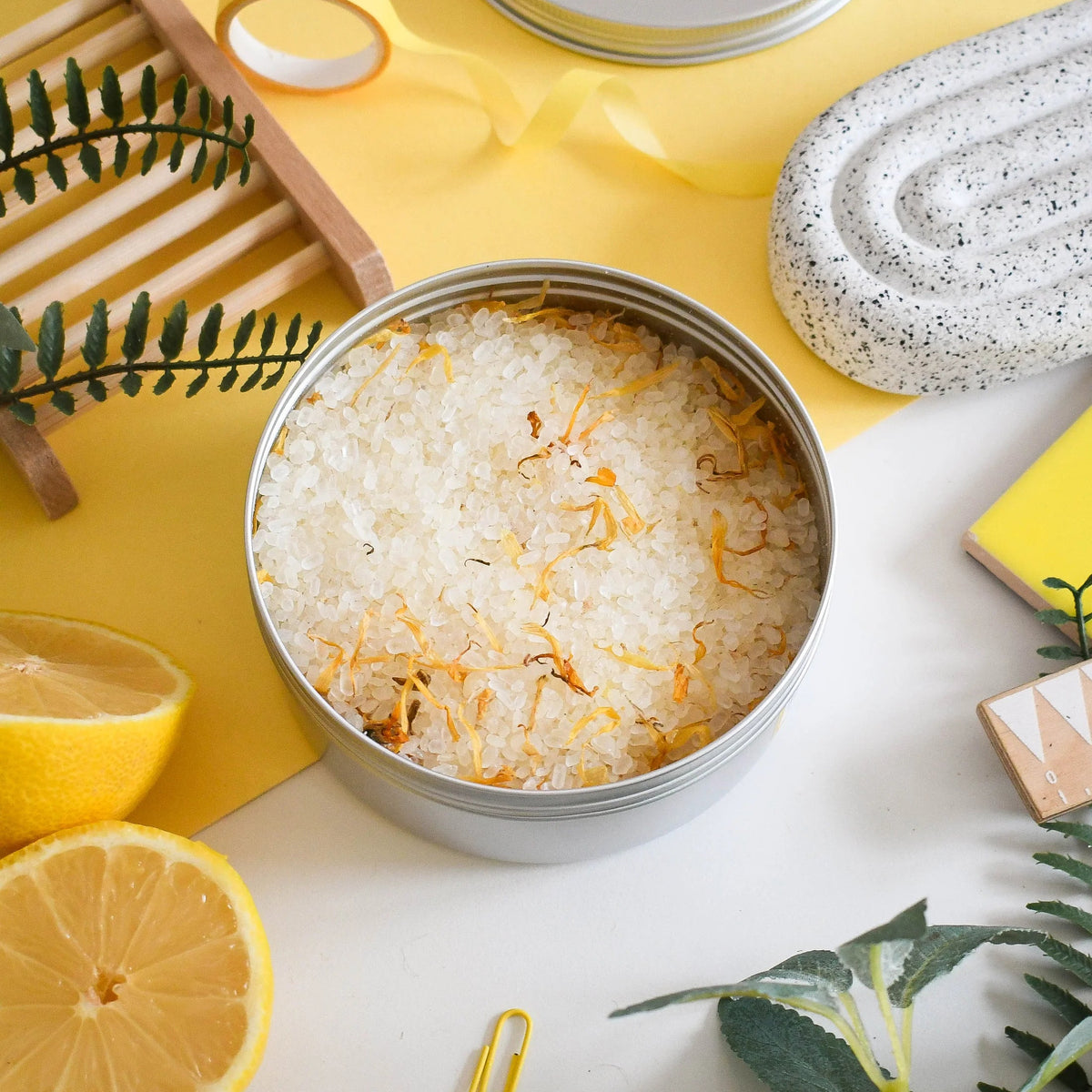 Metal tin filled with lemon bath salts on a yellow background with lemons and flowers.