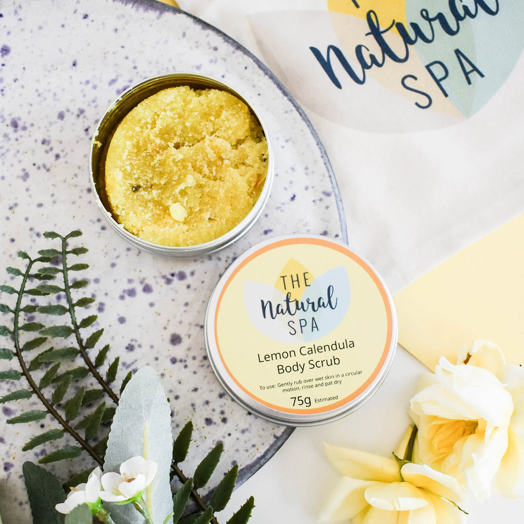 The Natural Spa Lemon Calendula Body Scrub with packaging on a decorative surface with flowers and leaves.