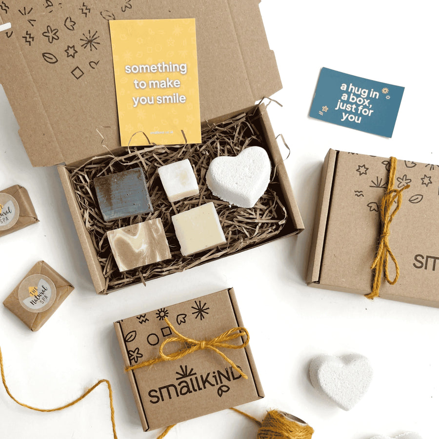 Little Luxuries Pamper box including mini soap, shampoo, conditioner and face wash bars along with a white heart shaped bath bomb. The products are inside a hand stamped kraft gift box filled with shredded paper and a yellow card which says 'something to make you smile’.