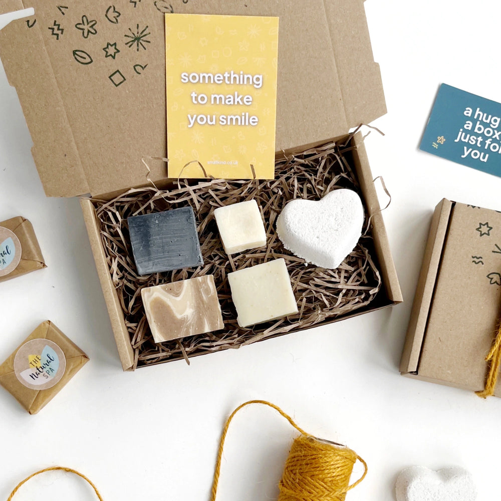 close up of the Smallkind Little Luxuries Pamper box including mini soap, shampoo, conditioner and face wash bars along with a white heart shaped bath bomb. The products are inside a kraft gift box filled with shredded paper and a yellow card which says 'something to make you smile’.