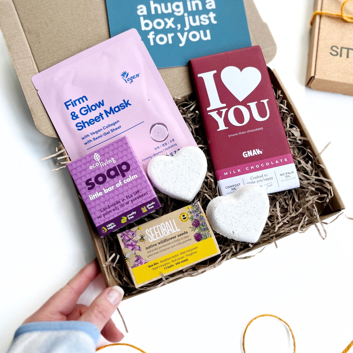 white womans hand holding the Love You Letterbox Gift: Eco-friendly pampering treats, including face mask, bath bombs, chocolate, and wildflower seeds