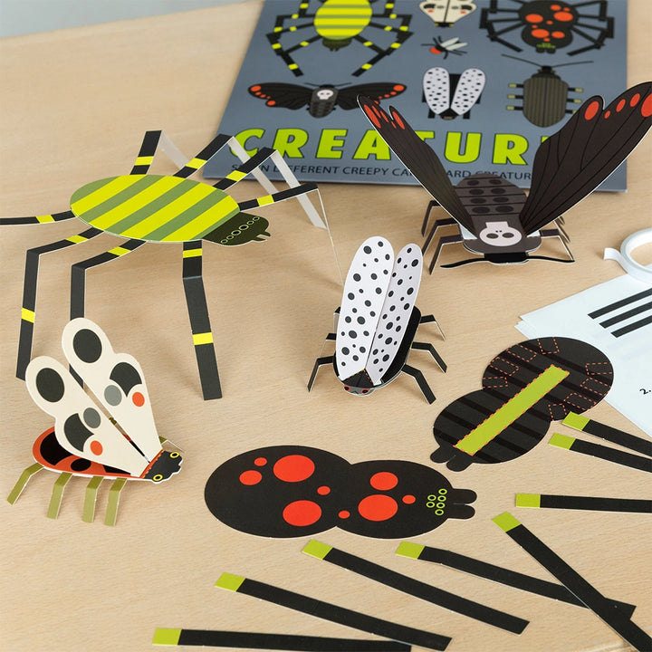 Collection of paper insect models on a wooden surface with a 'make Your Own Creepy Creatures' packaging in the background.
