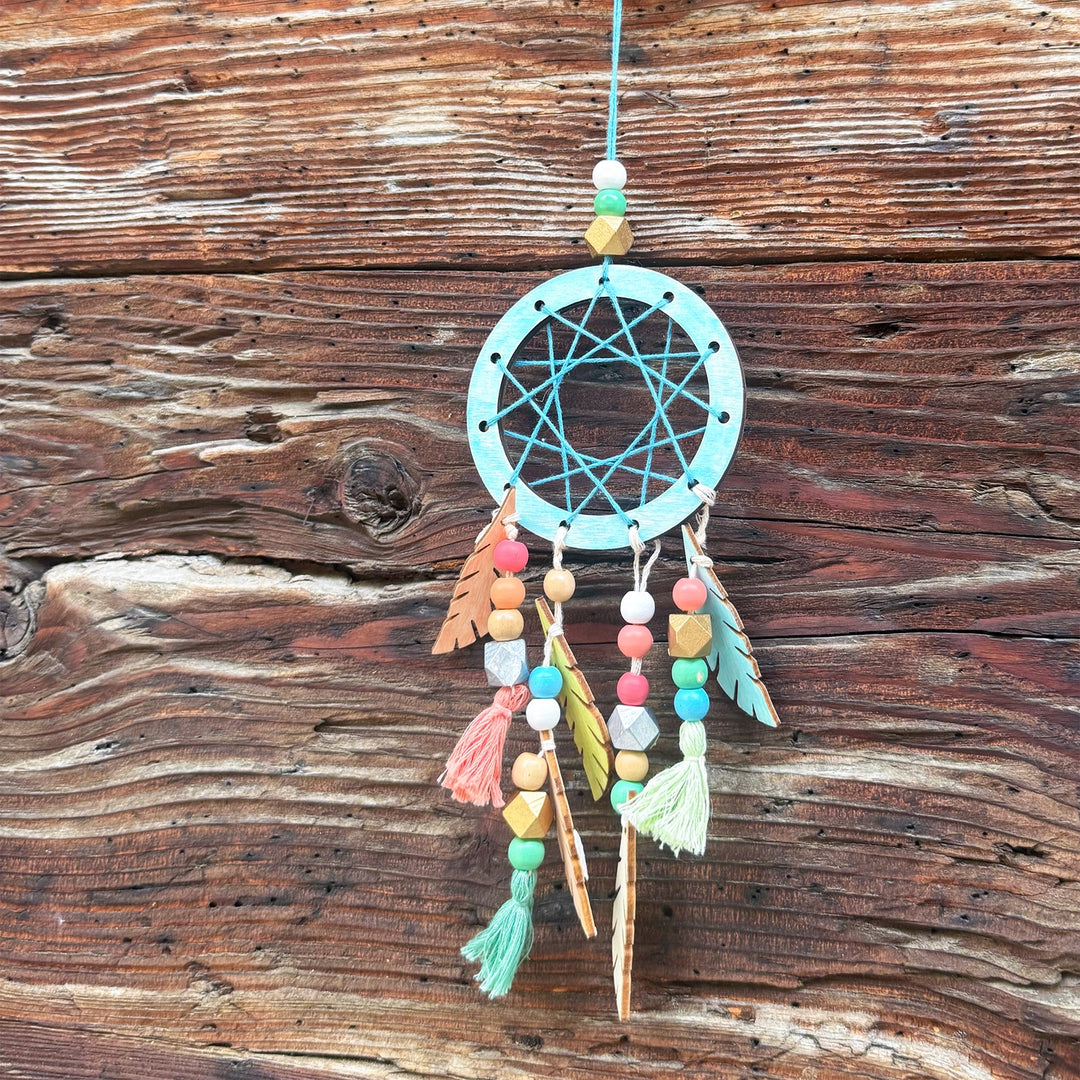 A colourful dreamcatcher with a light blue-green hoop, turquoise webbing, and multiple hanging tassels and feathers in pastel and natural tones, displayed against a rustic brown wooden plank background.